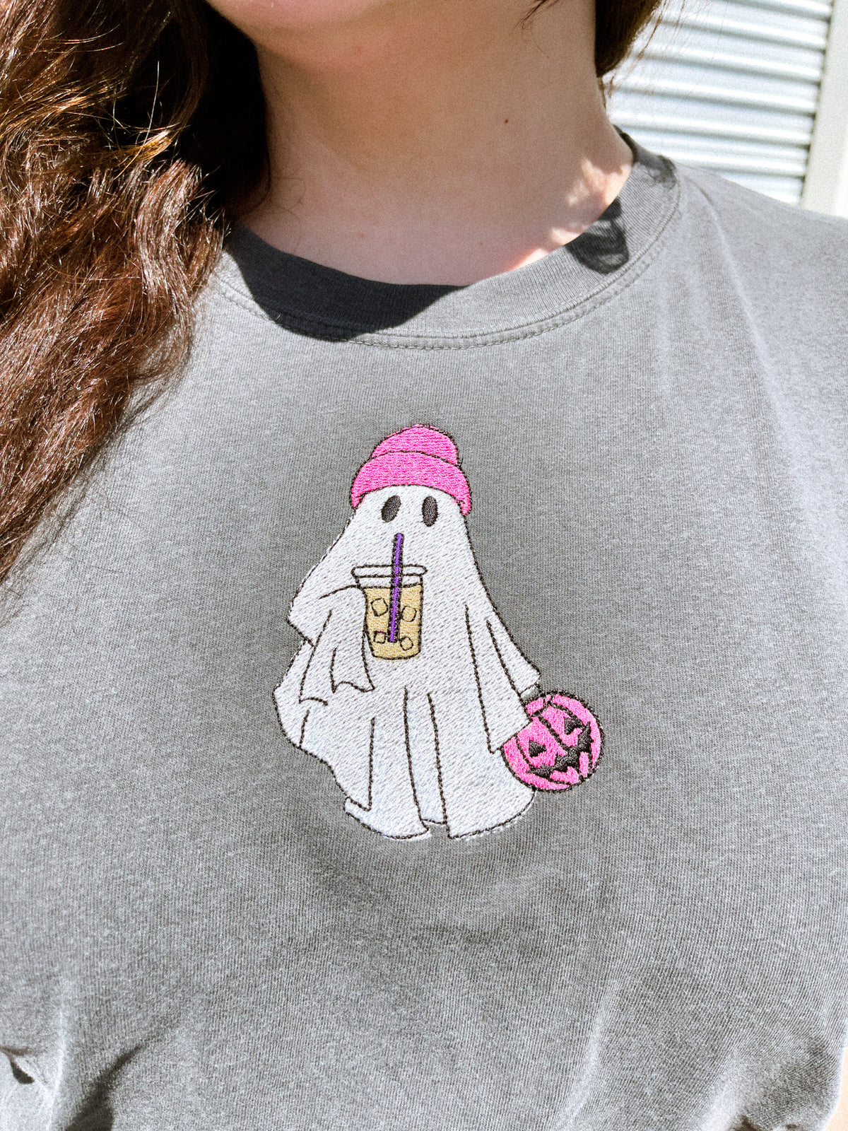 Up close picture of a embroidered pepper gray shirt with a white ghost design wearing a hot pink beanie, drinking an iced coffee, and carrying a hot pink jack-o-latern pumpkin